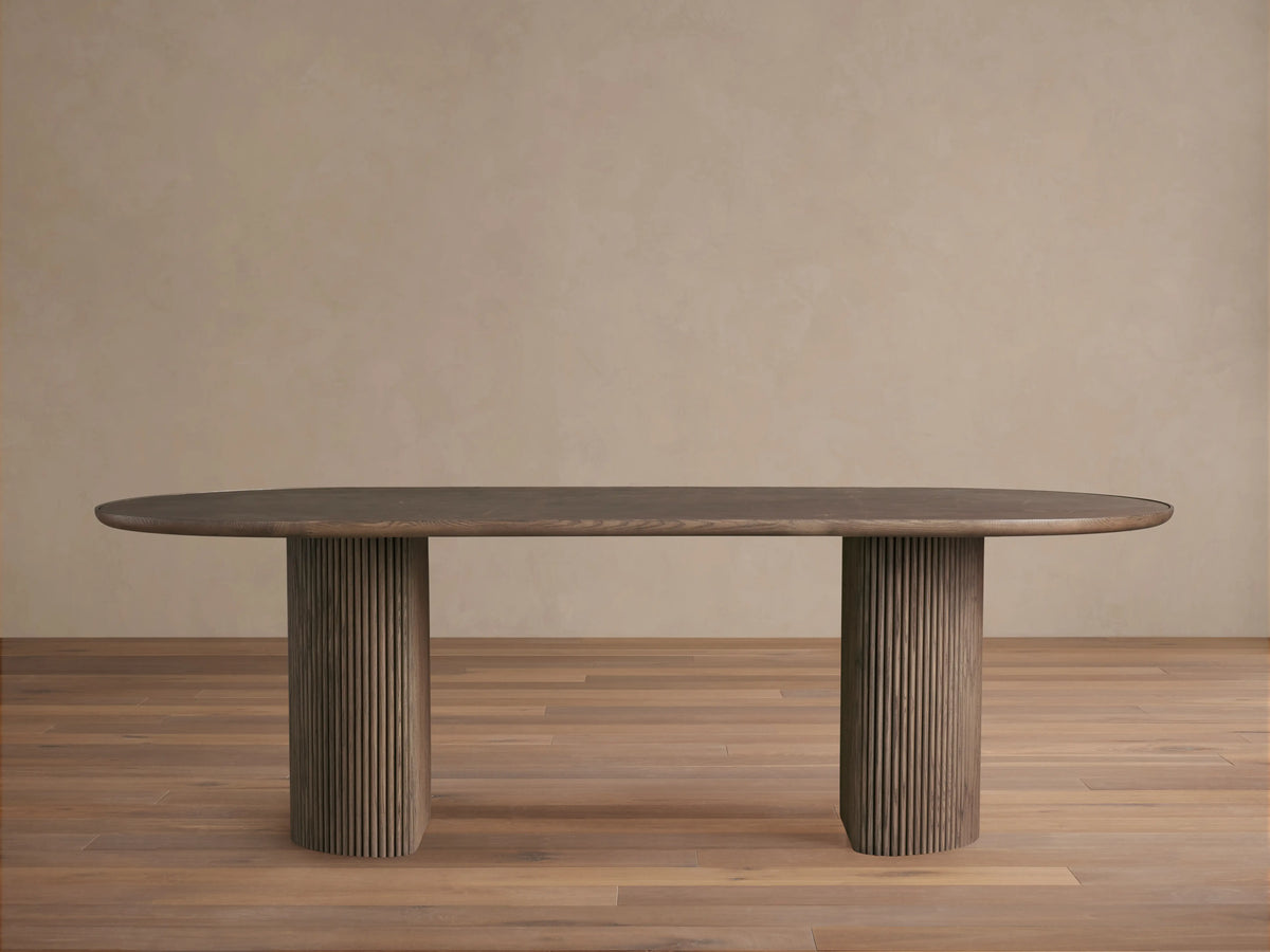 Astor Dining Table with elongated oval top in oak veneer, supported by two ribbed, cylindrical oak wood legs, on a wooden floor.