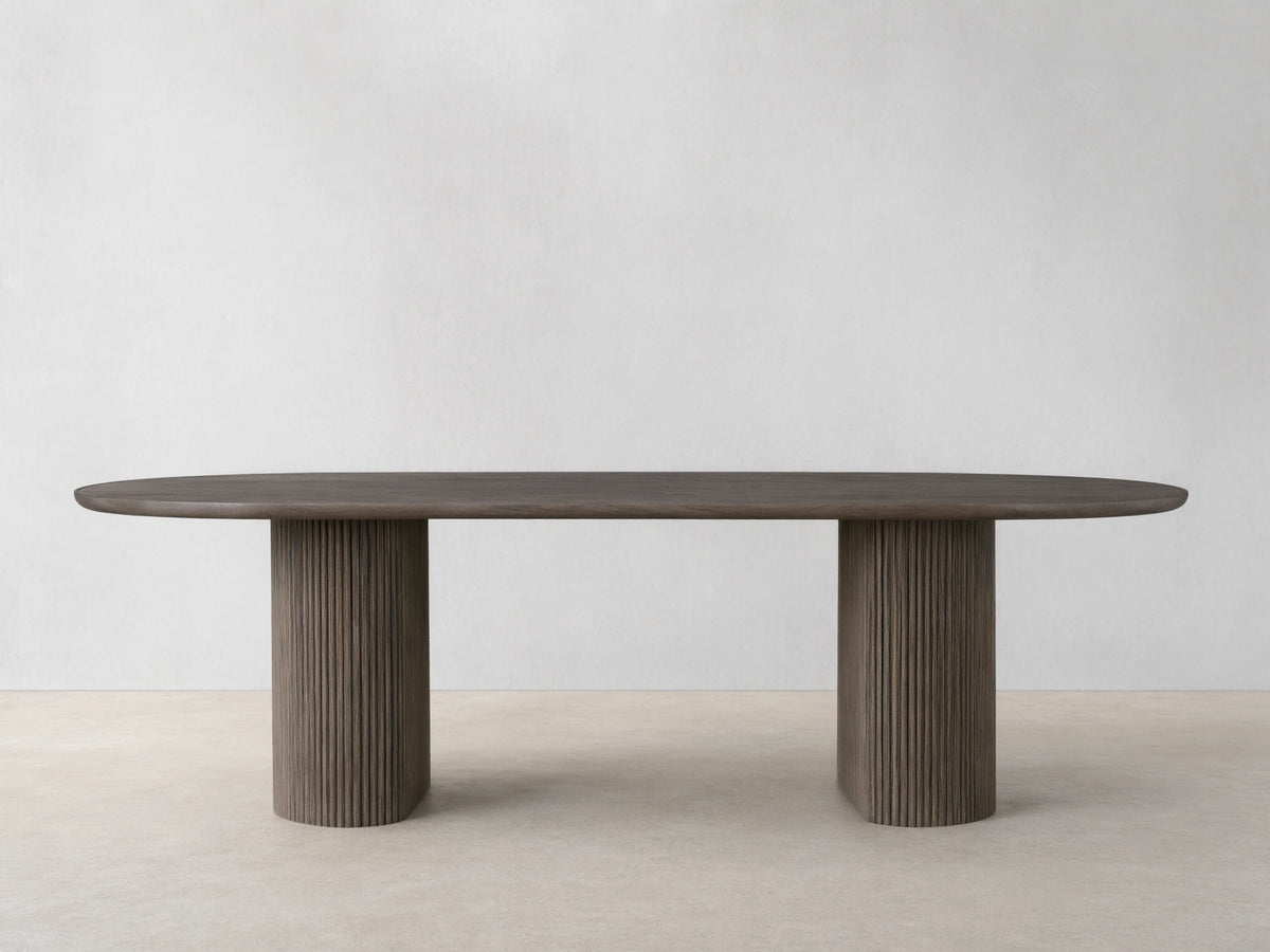 Astor Dining Table with elongated oval top in oak veneer, supported by two ribbed, cylindrical oak wood legs, on a wooden floor.