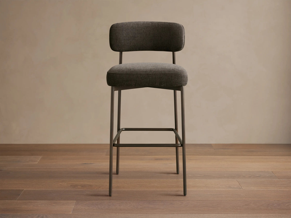 Billie Stool: Dark gray upholstered seat/back, antiqued dark iron frame, footrest. On wood floor.