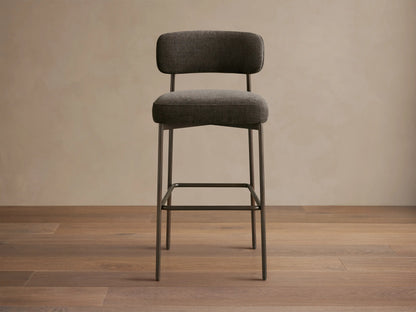 Billie Stool: Dark gray upholstered seat/back, antiqued dark iron frame, footrest. On wood floor.