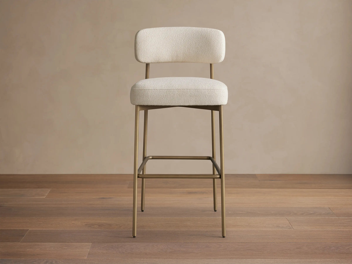 Billie Stool with off-white fabric cushioned seat & back, on antiqued gold iron frame, angled slightly right on wood floor.