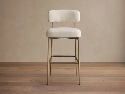 Billie Stool with off-white fabric cushioned seat & back, on antiqued gold iron frame, angled slightly right on wood floor.