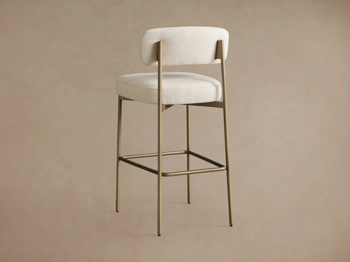 Close-up of Billie Stool: cream textured fabric upholstery, antiqued gold iron frame, showing rounded backrest and seat.