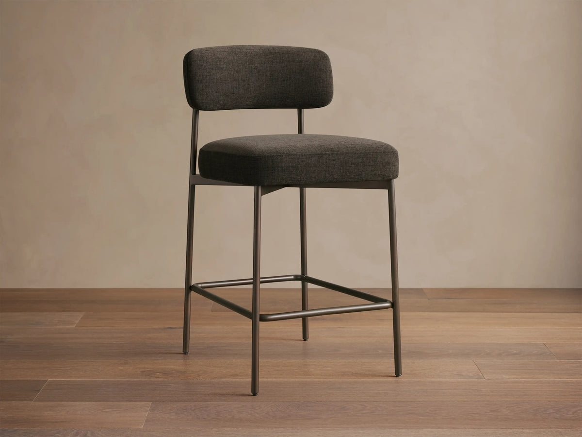 Billie Stool, dark gray fabric seat & back on antiqued iron frame, front-right view on a wooden floor.