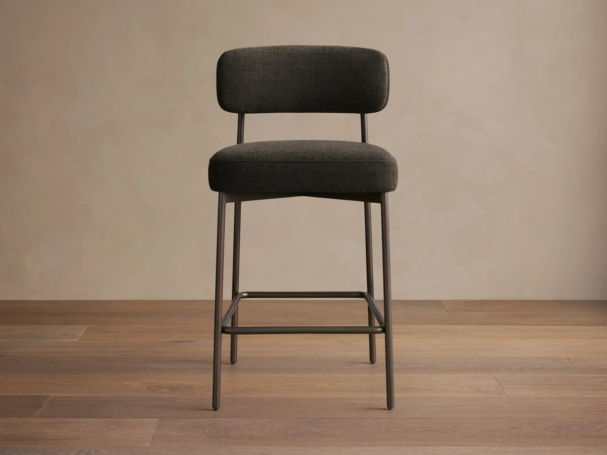 Billie Stool, dark gray fabric seat & back on antiqued iron frame, front-right view on a wooden floor.