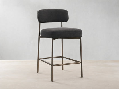 Billie Stool, dark gray fabric seat & back on antiqued iron frame, front-right view on a wooden floor.