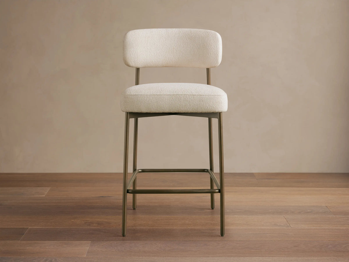 Billie Stool: Cream textured fabric seat & back on an antiqued bronze metal frame, with a footrest, on a wooden floor.