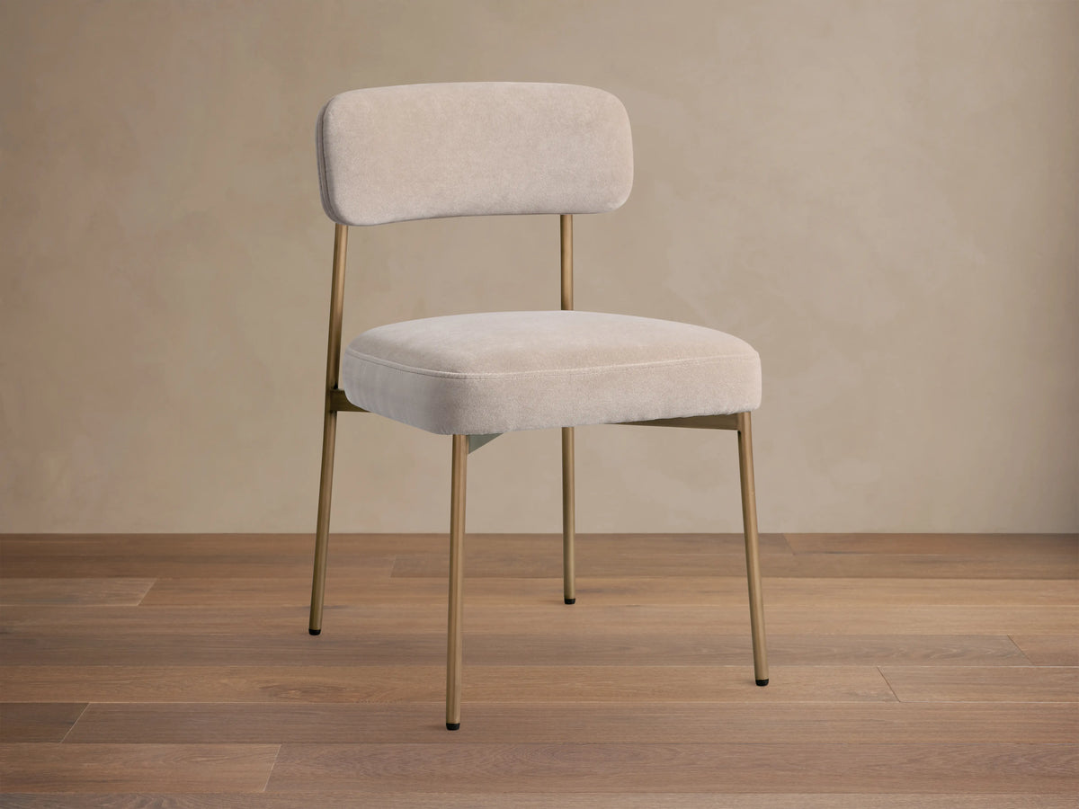Front 3/4 view of a beige upholstered dining chair, featuring a curved back, soft cushion, and thin gold-toned metal legs.