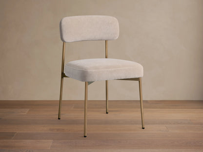 Front 3/4 view of a beige upholstered dining chair, featuring a curved back, soft cushion, and thin gold-toned metal legs.