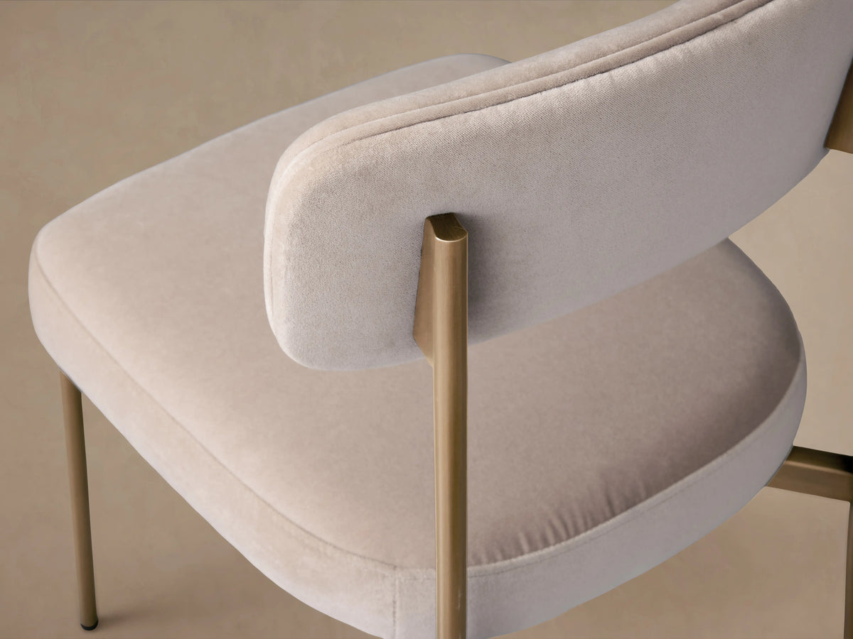Angled close-up of a cream velvet chair with a curved backrest and golden metal frame.