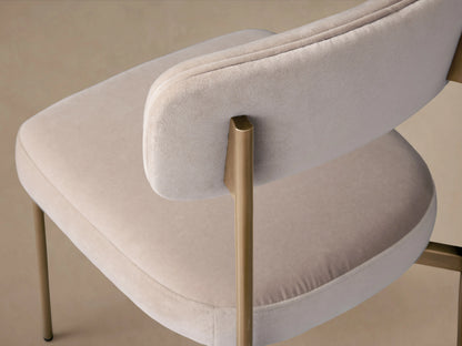 Angled close-up of a cream velvet chair with a curved backrest and golden metal frame.
