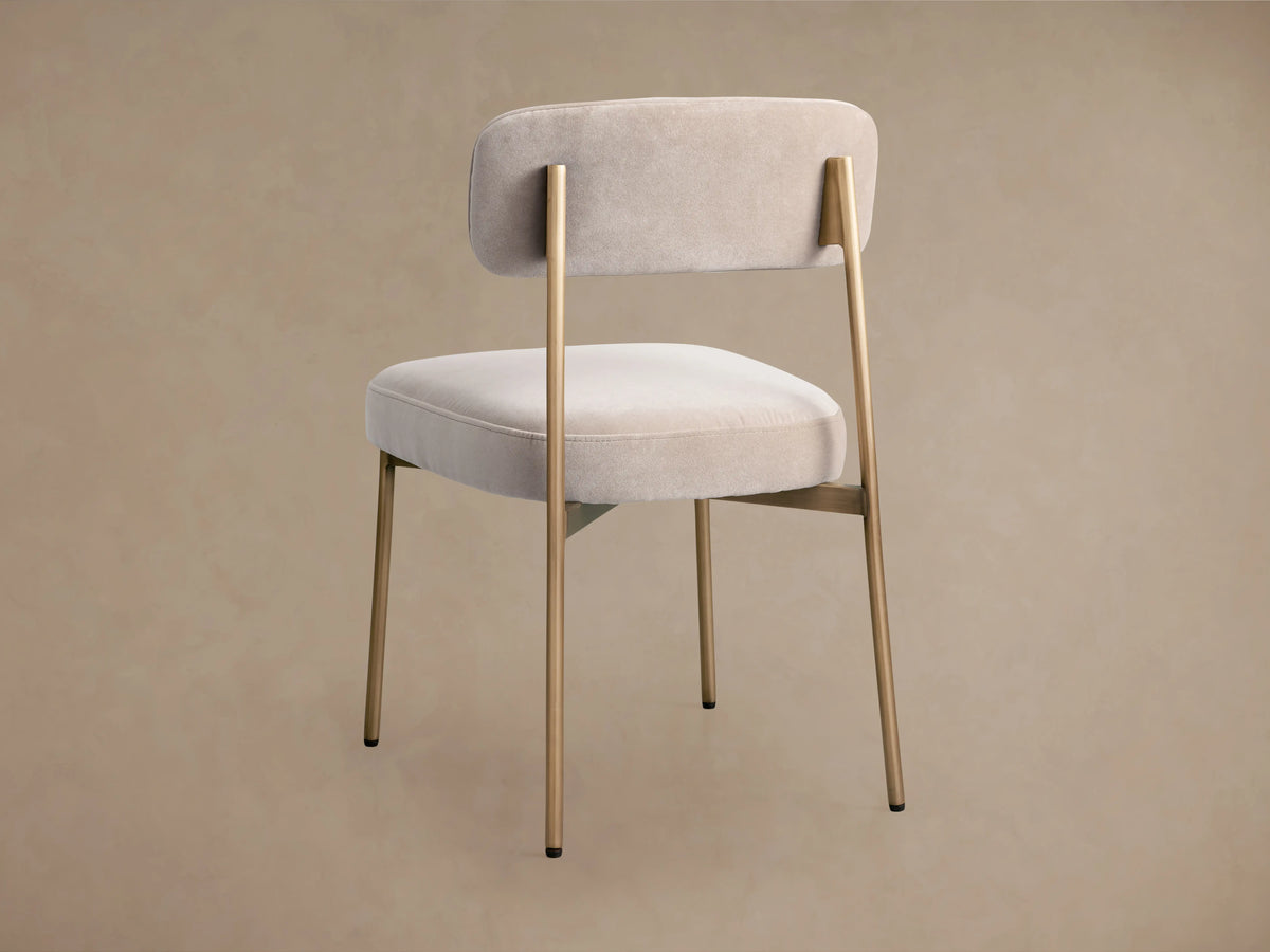 Rear view of a beige upholstered chair with a rounded backrest and seat, supported by thin gold metal legs.