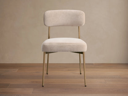 Front view of a beige velvet dining chair, featuring a rounded backrest and seat, with slender golden metal legs.