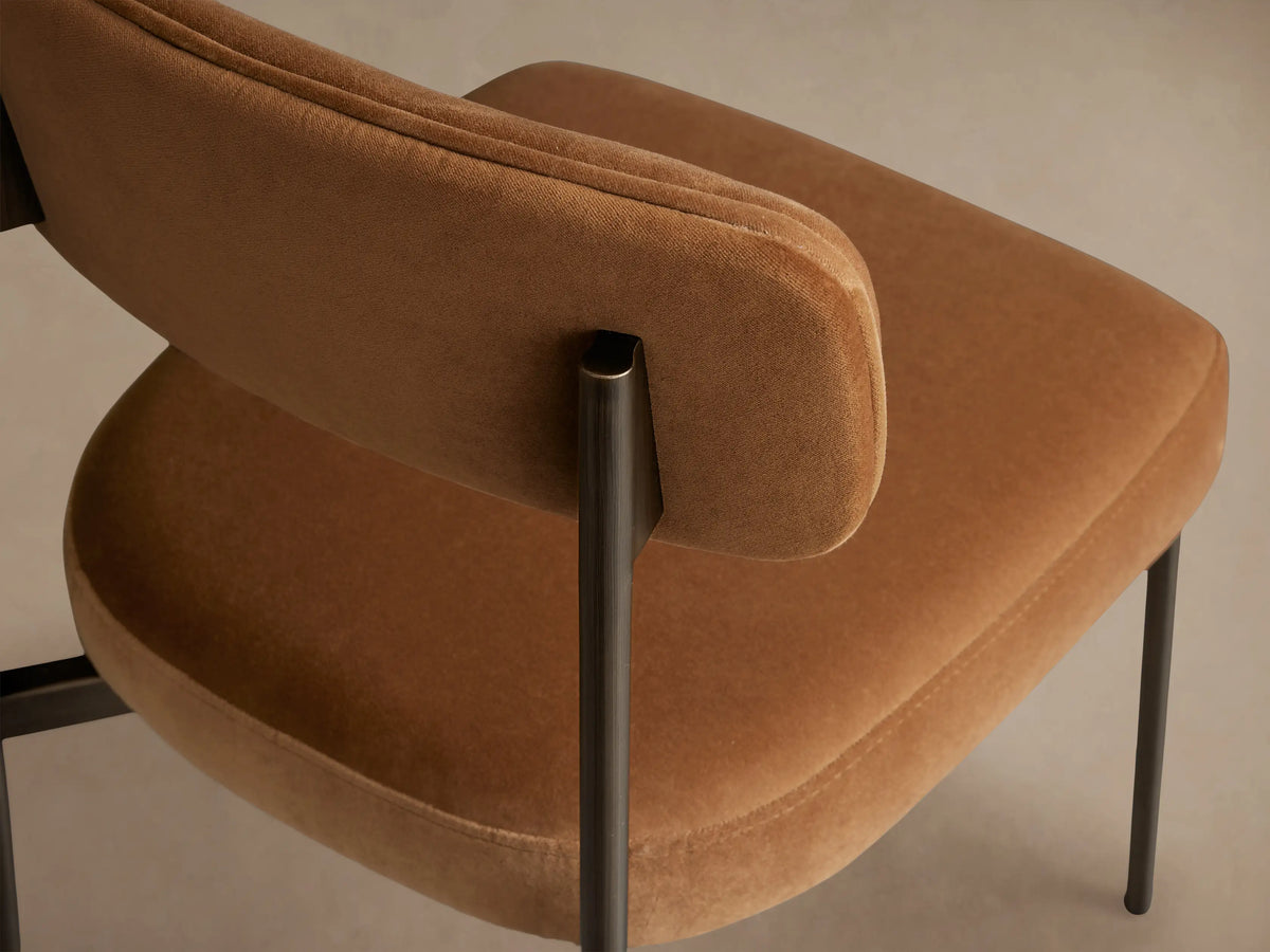 Billie Dining Chair, front. Rounded brown upholstered seat & back, antique bronze frame & legs, on wood floor, light wall.