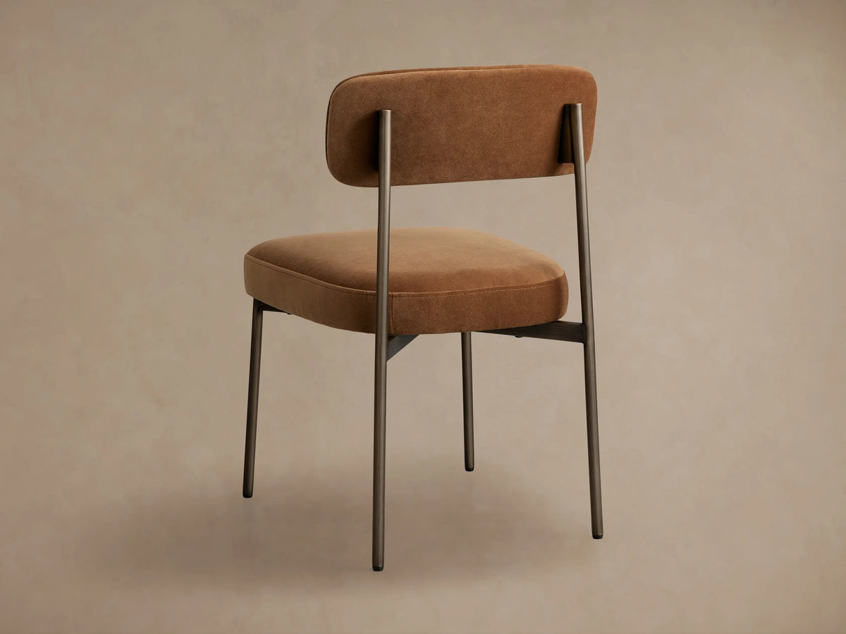 Angled close-up of Billie Dining Chair. Curved back and seat in soft, caramel-brown fabric, dark antique bronze iron frame.
