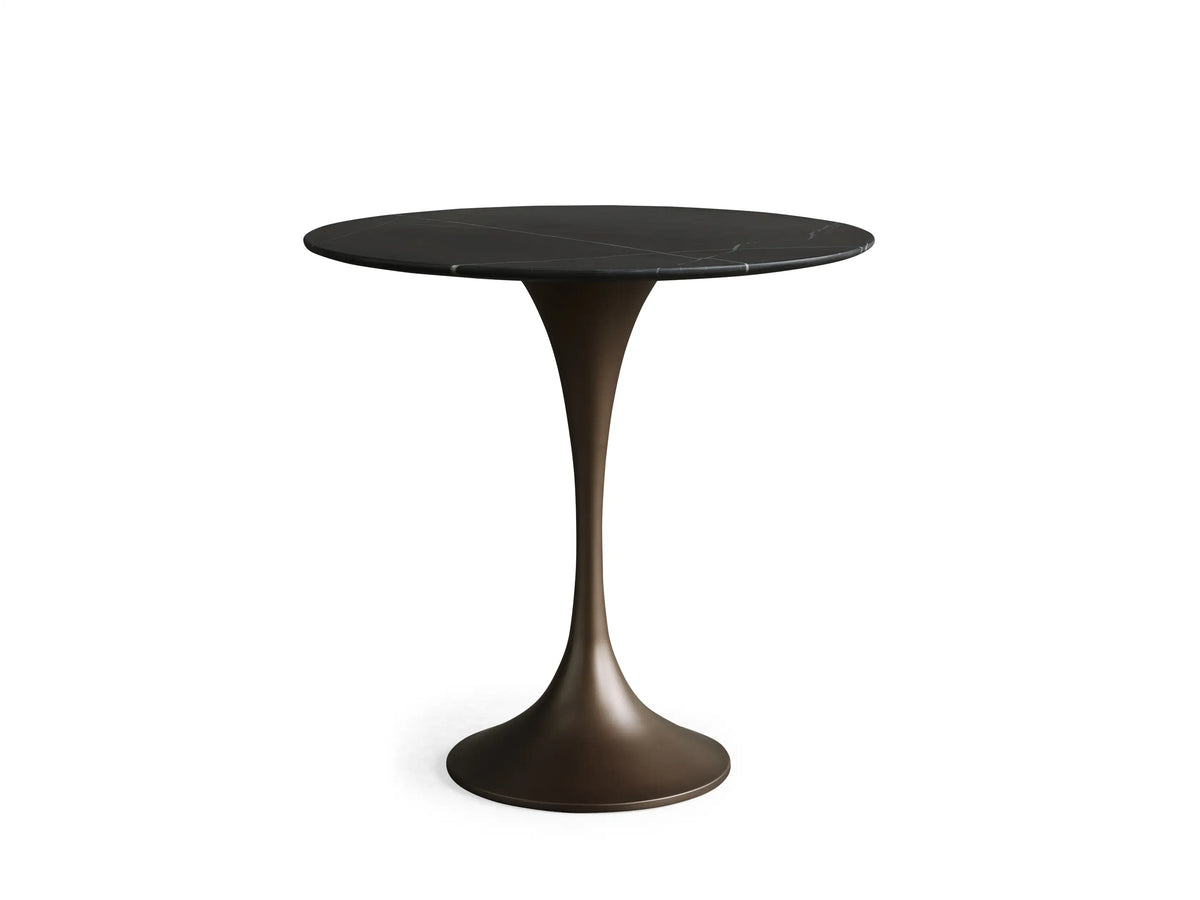 Saint Germain Bistro Table with a black round stone tabletop and sleek bronze steel base, on a wooden floor against a beige wall.