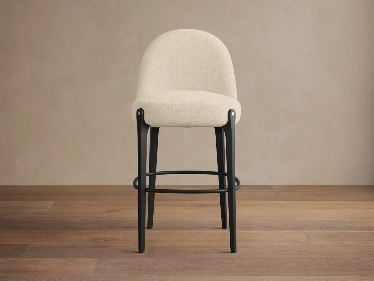 Bottoni Stool with a cream upholstered seat and curved back, black legs, metal footrest, and brass tacks, angled slightly right.