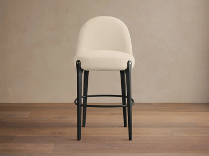 Bottoni Stool with a cream upholstered seat and curved back, black legs, metal footrest, and brass tacks, angled slightly right.