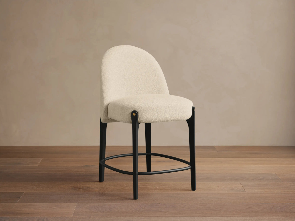 Bottoni Stool: cream textured upholstery, rounded back, dark wood legs with brass tacks, and a metal footrest ring.