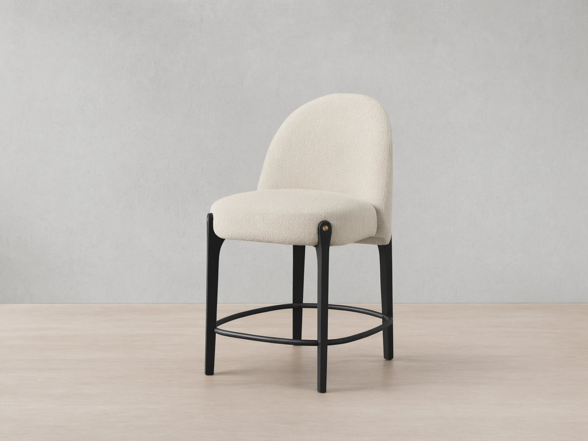 Bottoni Stool: cream textured upholstery, rounded back, dark wood legs with brass tacks, and a metal footrest ring.