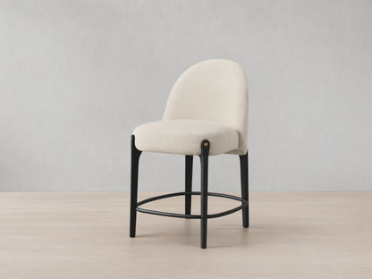 Bottoni Stool: cream textured upholstery, rounded back, dark wood legs with brass tacks, and a metal footrest ring.