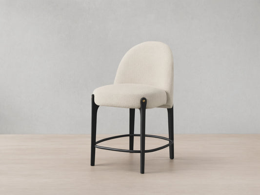 Bottoni Stool: cream textured upholstery, rounded back, dark wood legs with brass tacks, and a metal footrest ring.