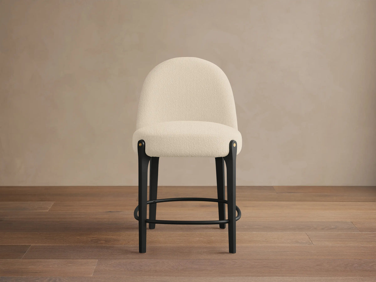 Bottoni Stool: cream textured upholstery, rounded back, dark wood legs with brass tacks, and a metal footrest ring.