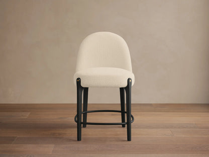 Bottoni Stool: cream textured upholstery, rounded back, dark wood legs with brass tacks, and a metal footrest ring.