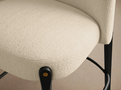 Bottoni Stool: a curved, cream textured upholstered seat and back, supported by a dark wood frame with brass accents and a metal footrest.