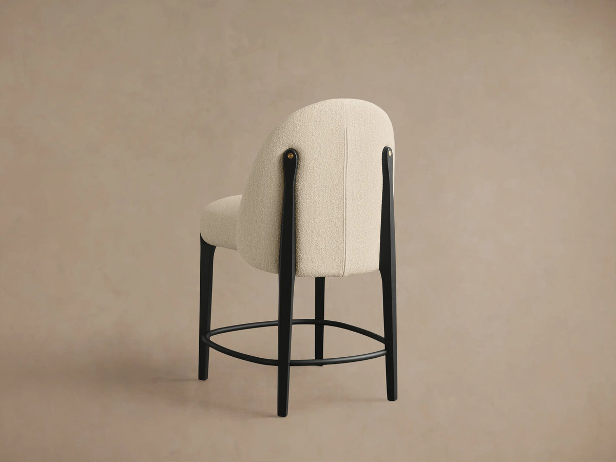 Close-up of Bottoni Stool's cream bouclÃ© upholstered seat and back, with black leg and brass accent on light beige background.