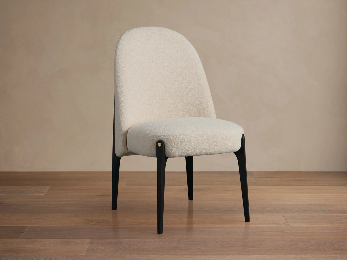 Off-white boucle Bottoni Dining Chair with rounded back, dark tapered legs, and brass accent, on a wood floor.