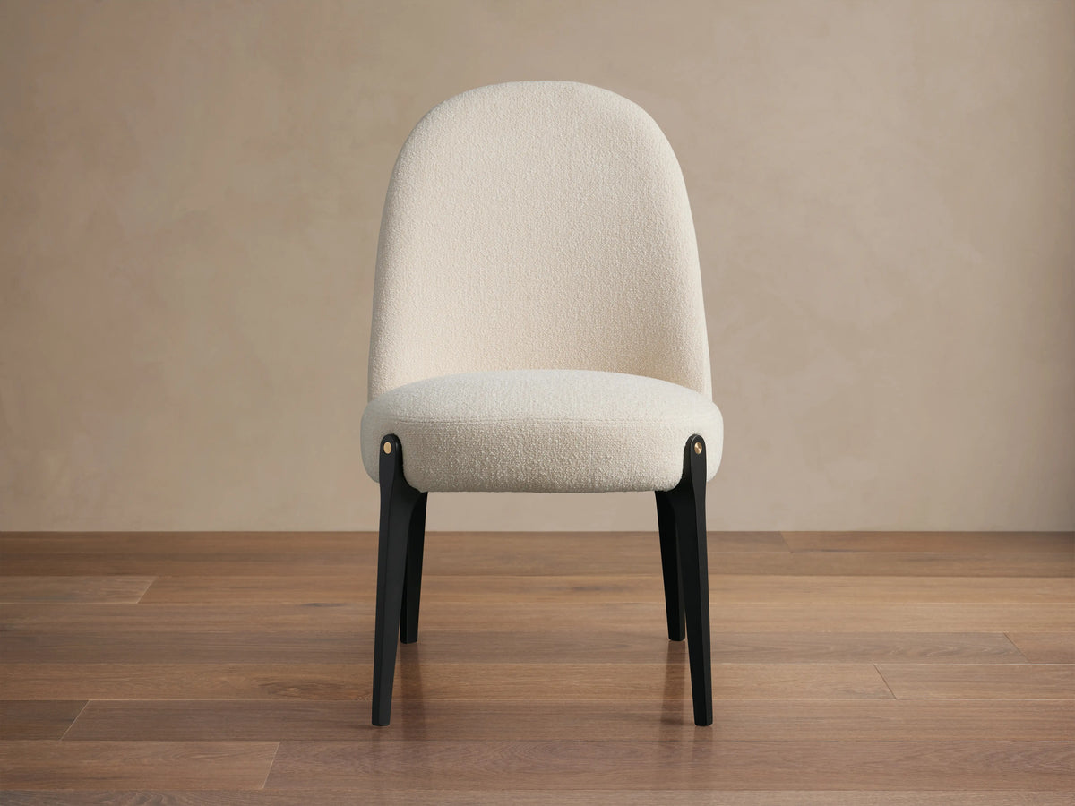Off-white boucle Bottoni Dining Chair with rounded back, dark tapered legs, and brass accent, on a wood floor.
