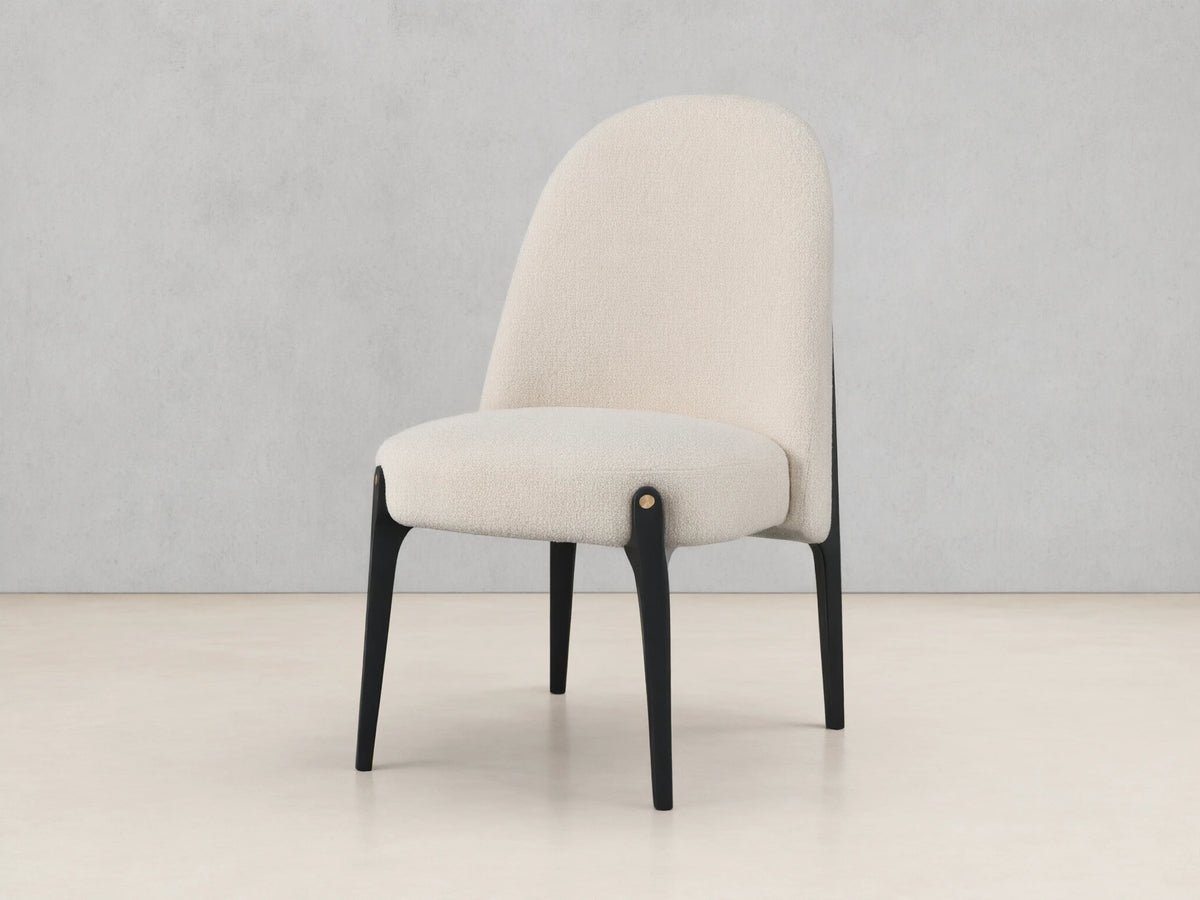 Off-white boucle Bottoni Dining Chair with rounded back, dark tapered legs, and brass accent, on a wood floor.