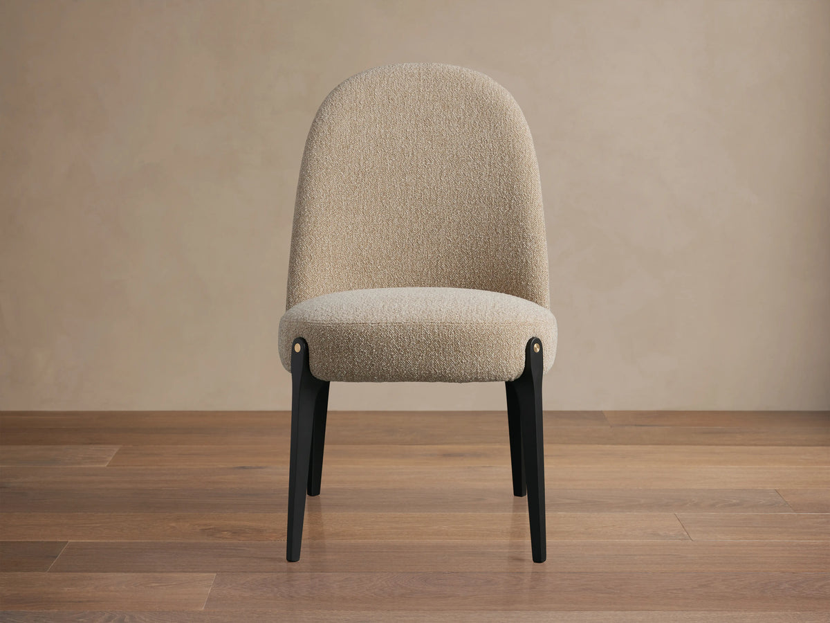 Bottoni Dining Chair with light beige textured upholstery, dark legs, and brass accents, angled slightly right on a wood floor.