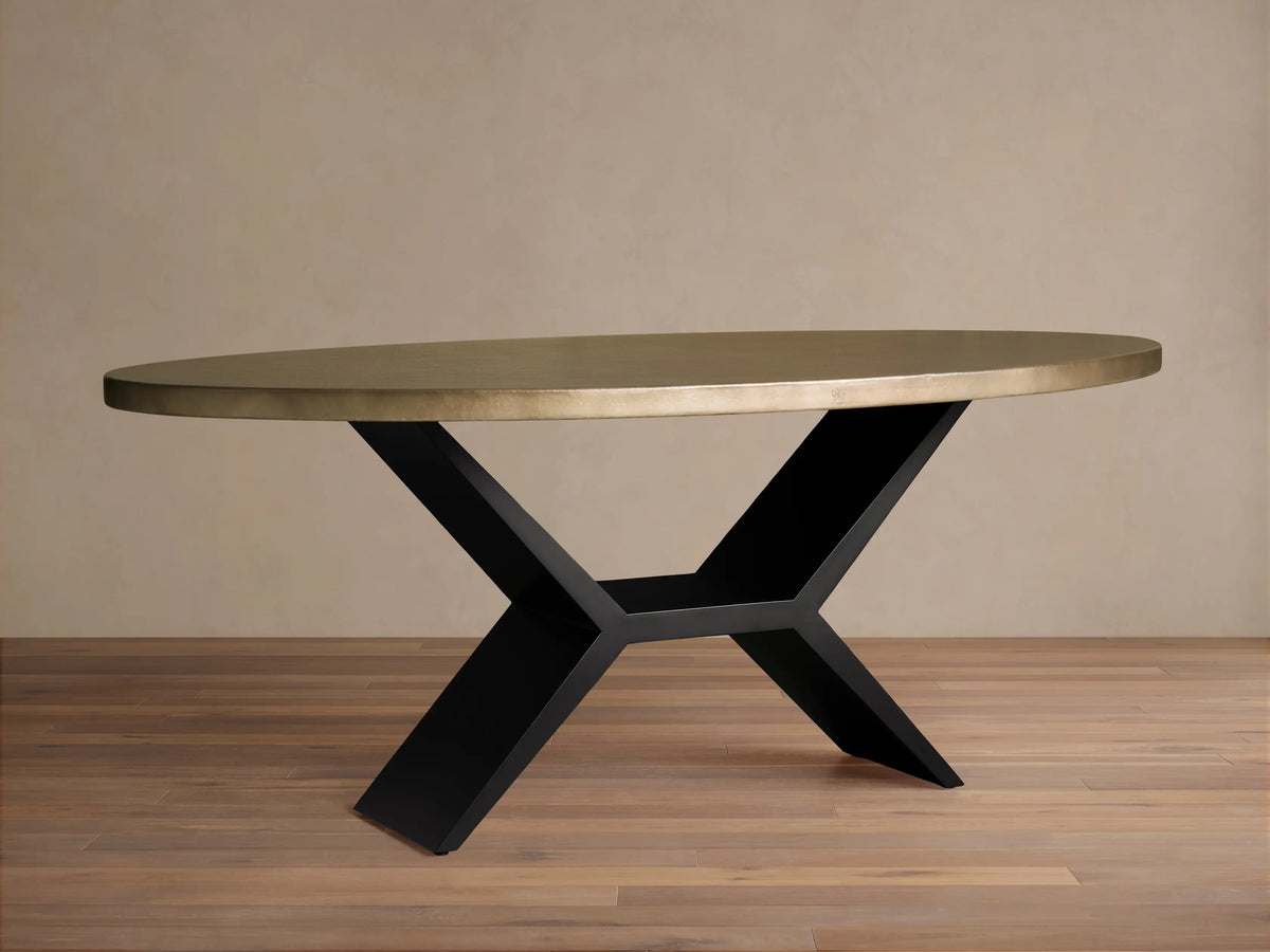 Oval dining table with a brass-toned recycled metal top and a striking black iron Vertex Base, against a neutral wall.
