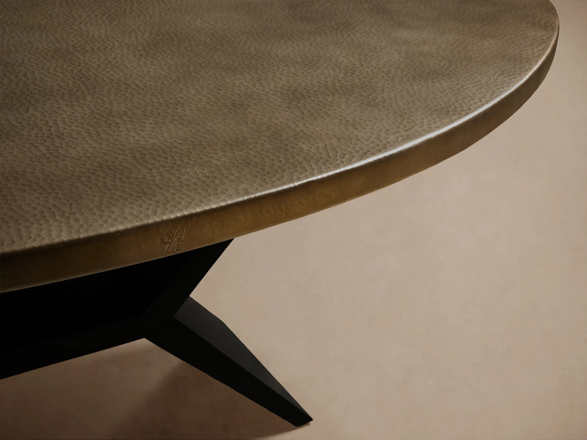 Recycled Metal Dining Table with an oval, brass-toned top and a black X-shaped Vertex base, on a wooden floor.