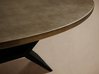 Recycled Metal Dining Table with an oval, brass-toned top and a black X-shaped Vertex base, on a wooden floor.