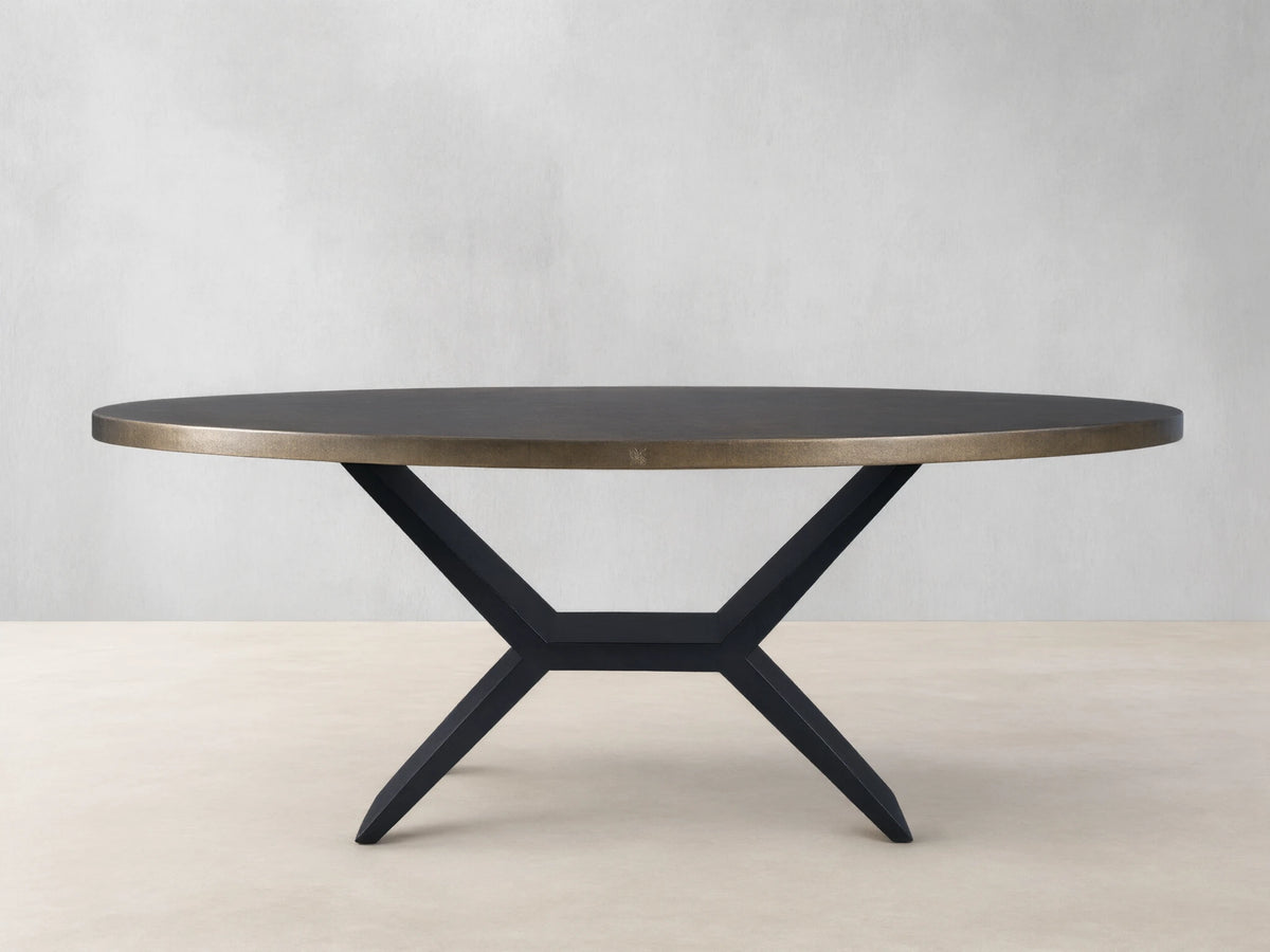 Oval dining table with a brass-toned recycled metal top and a striking black iron Vertex Base, against a neutral wall.