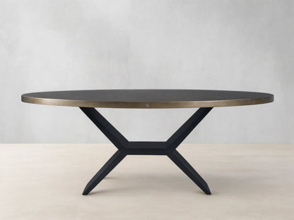 Oval dining table with a brass-toned recycled metal top and a striking black iron Vertex Base, against a neutral wall.
