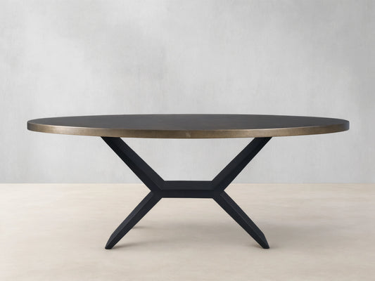 Oval dining table with a brass-toned recycled metal top and a striking black iron Vertex Base, against a neutral wall.