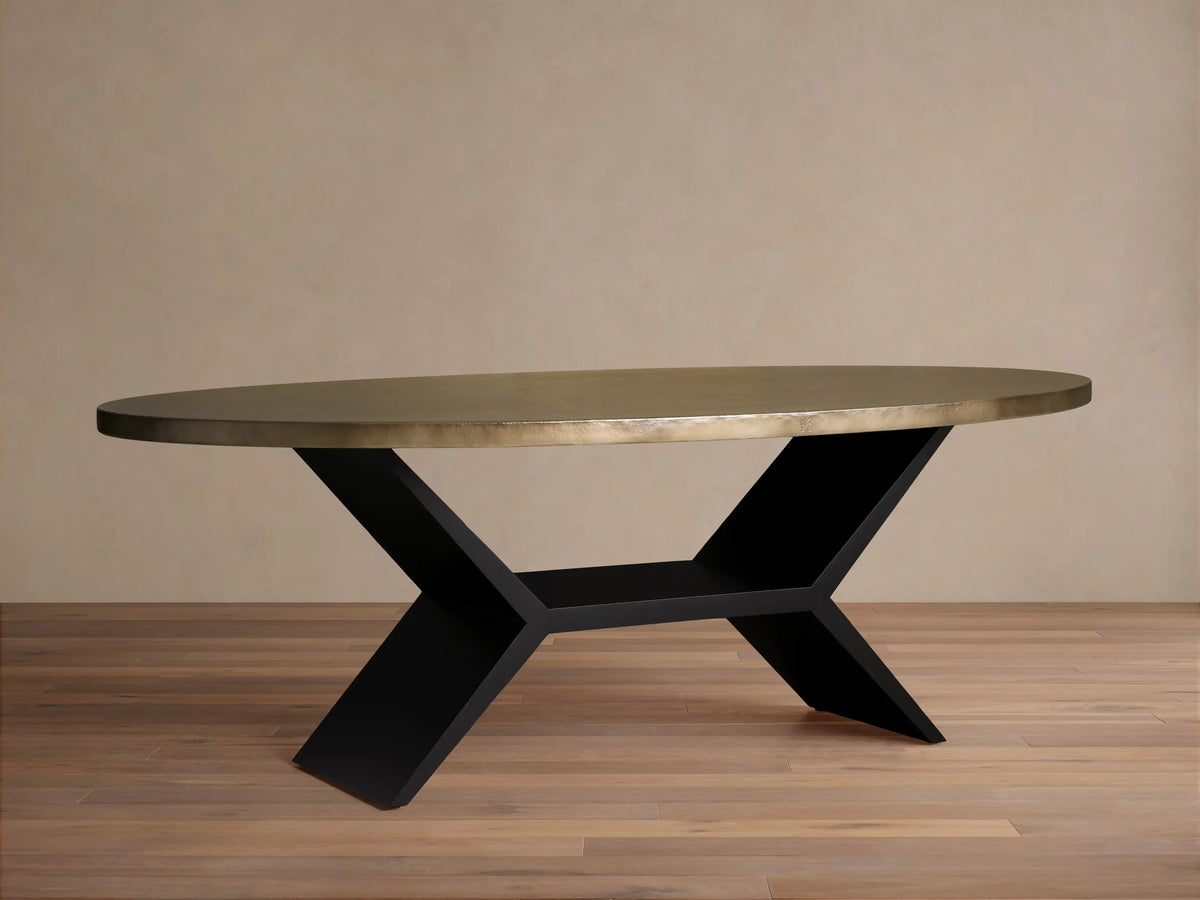 Oval recycled brass dining table with textured surface, on a geometric black Vertex base. Neutral wall, wood floor background.