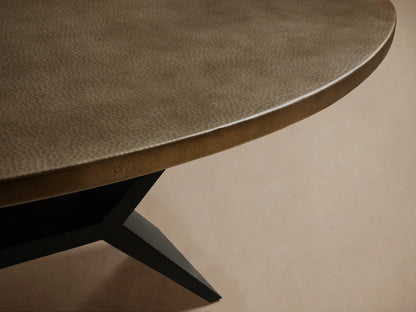 Recycled Metal Dining Table with an oval, warm brass-toned top & angular black Vertex Base on wood floor.