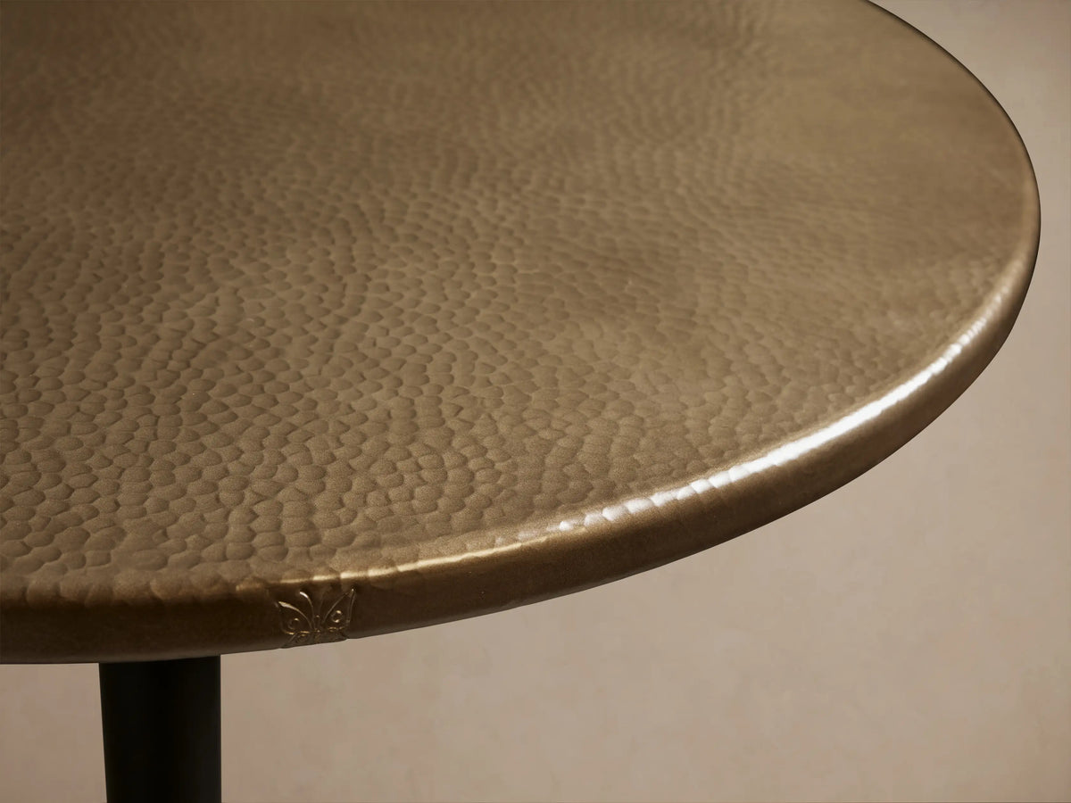 Round brass-finished recycled metal counter table with a single pedestal Wade iron base, centered on a wood floor.