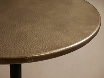 Round brass-finished recycled metal counter table with a single pedestal Wade iron base, centered on a wood floor.