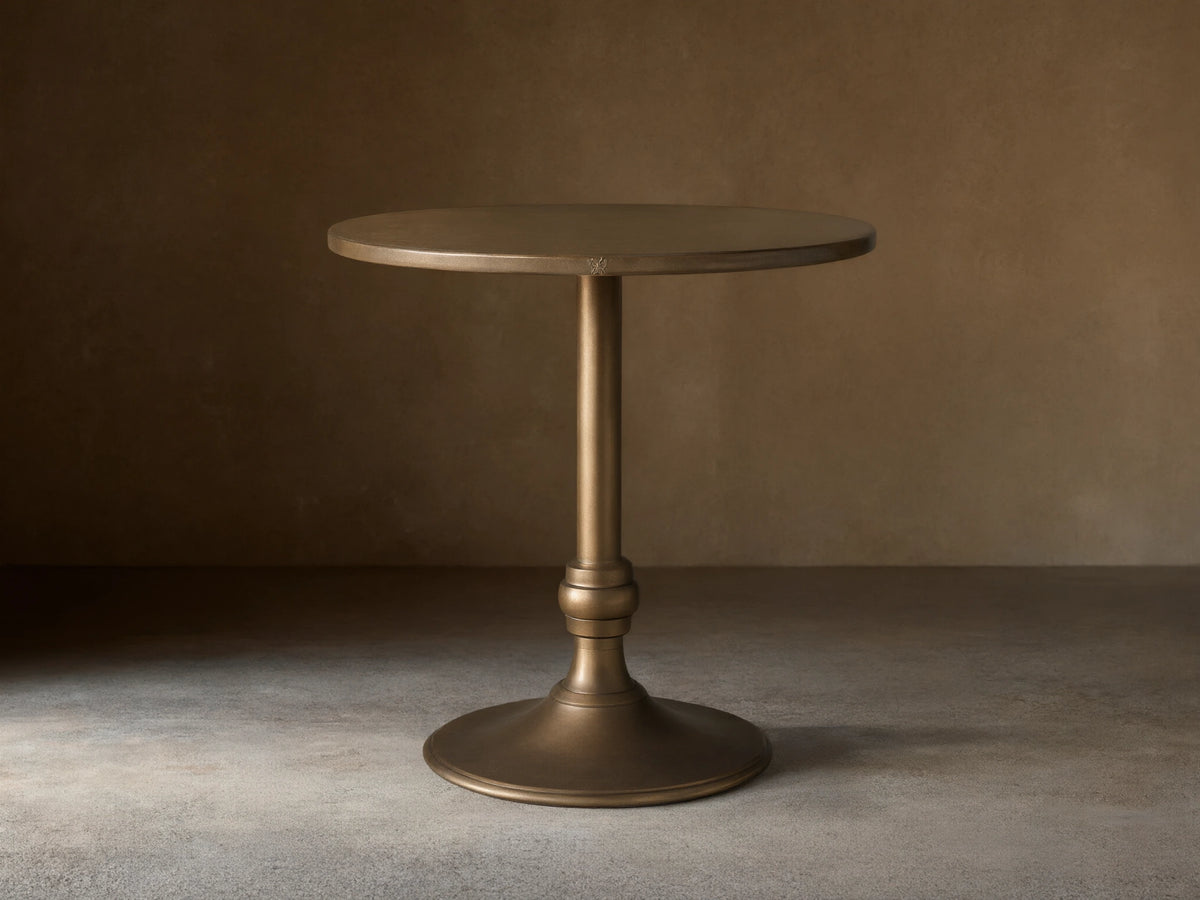 Round brass-finished recycled metal counter table with a single pedestal Wade iron base, centered on a wood floor.