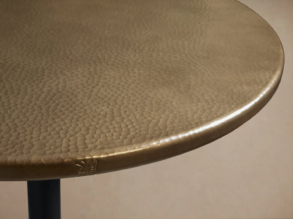 Recycled Metal Bar Table with a round, brass-finished top and a black, ornate Wade iron base on a wooden floor.