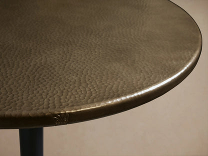 Recycled Metal Bistro Table with round brass-finished top and a dark Wade pedestal base, against a tan wall and wood floor.