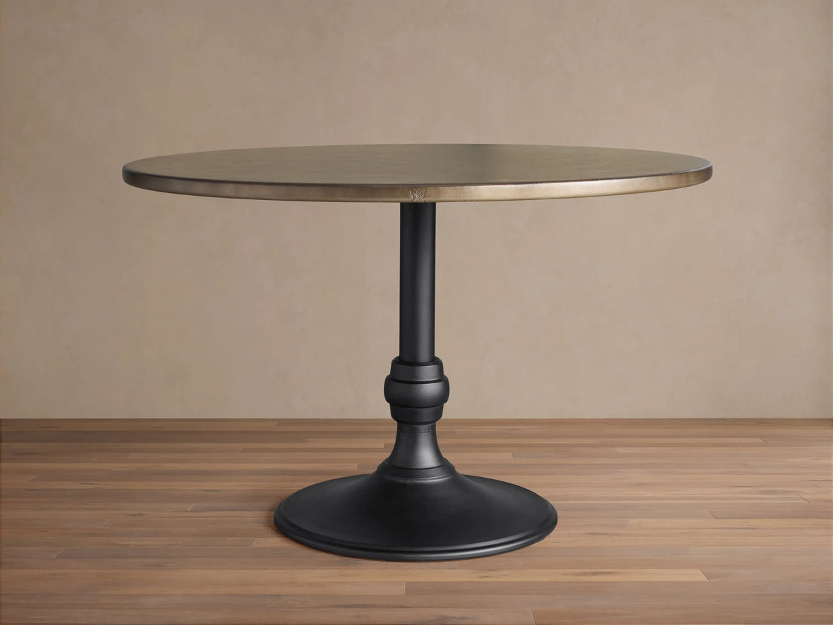 Recycled Metal Dining Table with Wade Base. Round bronze metal top, black ornate pedestal base on wooden floor.