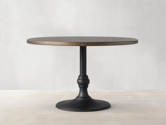 Recycled Metal Dining Table with Wade Base. Round bronze metal top, black ornate pedestal base on wooden floor.