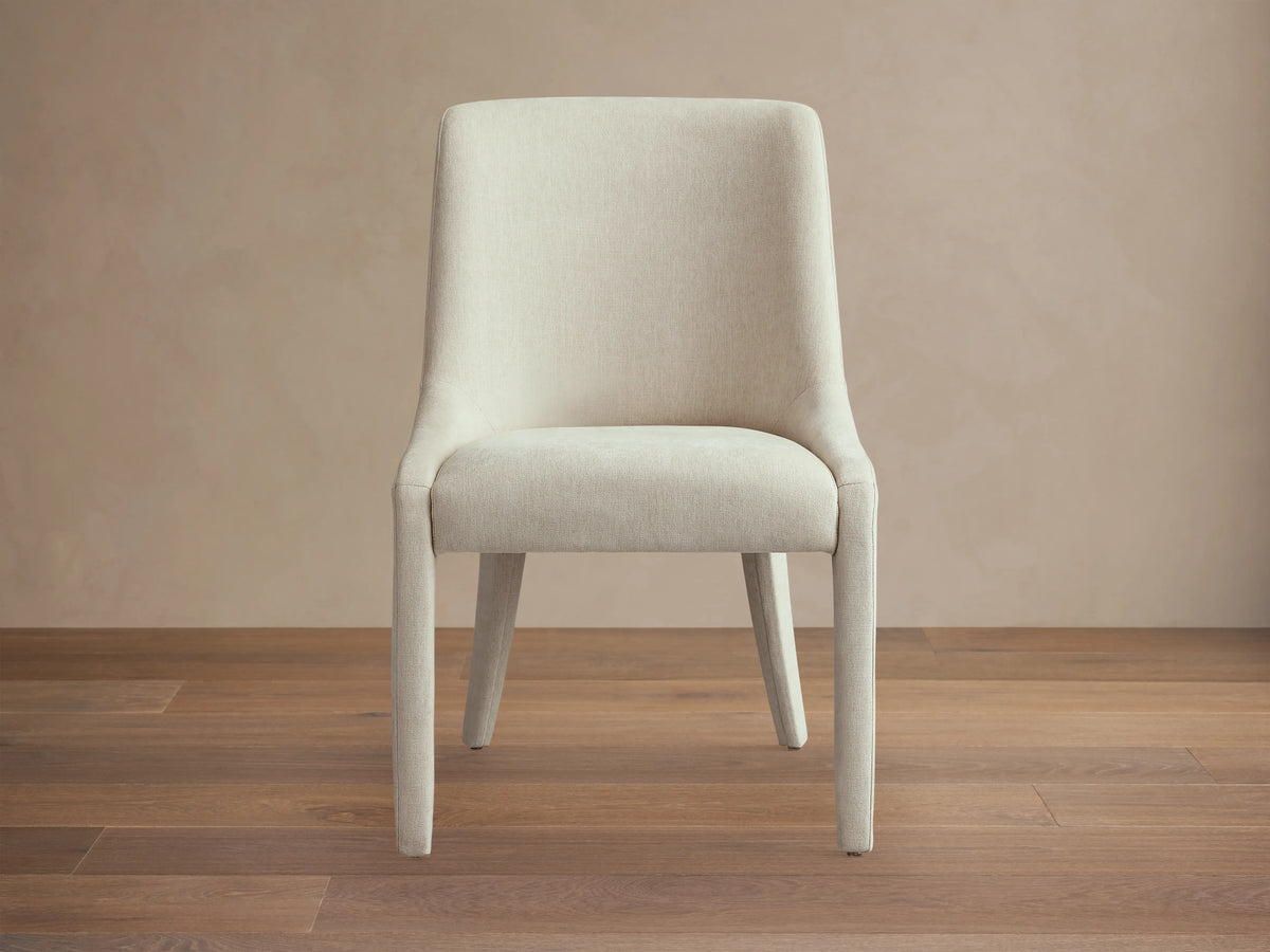 Burns Dining Chair in beige upholstery, with a rounded backrest and cushioned seat, featuring smooth, sloping lines on a plain background.